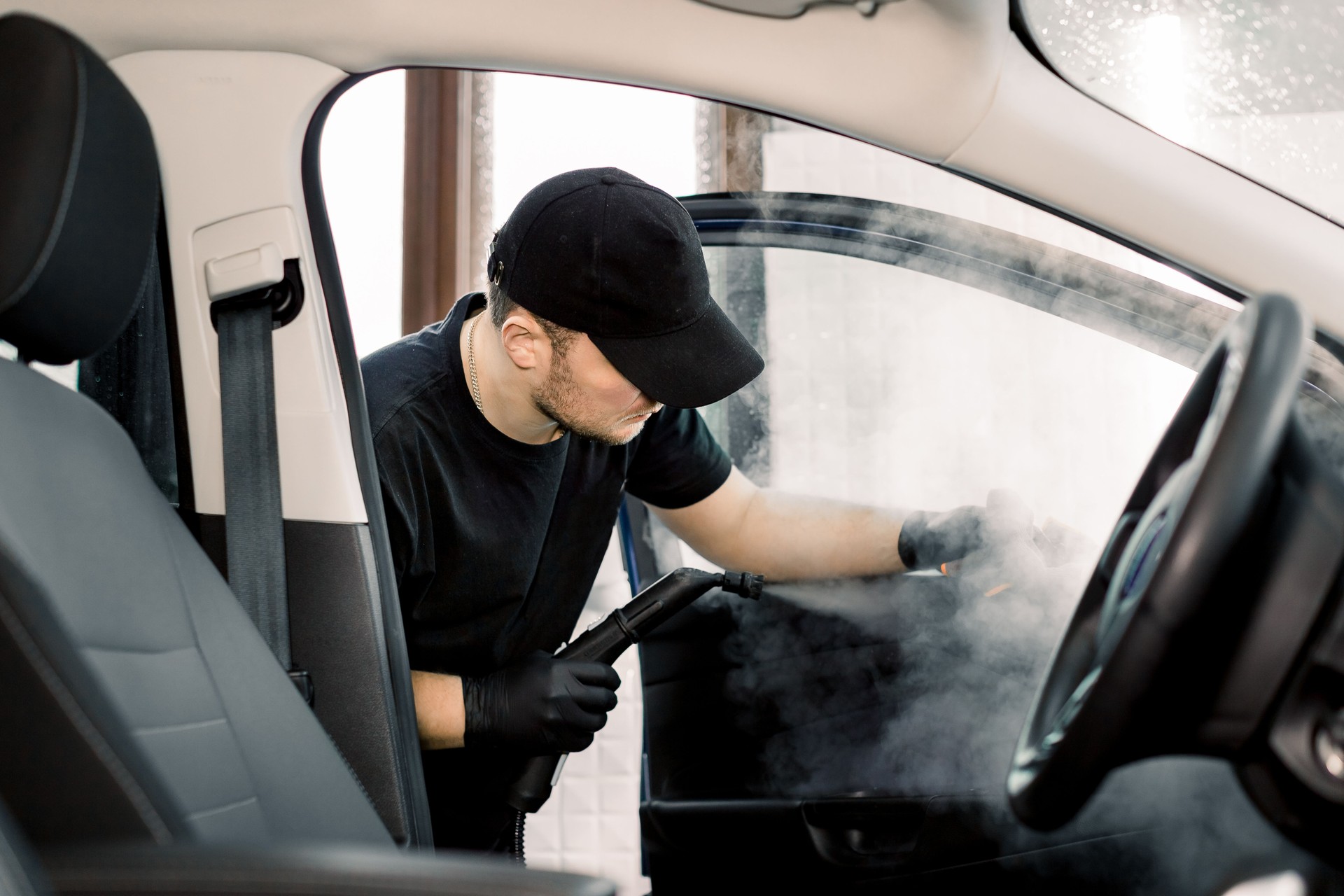 Auto cleaning service and detailing concept. Handsome Caucasian man in black uniform cleaning interior of the car with hot steam cleaner. Selective focus.
